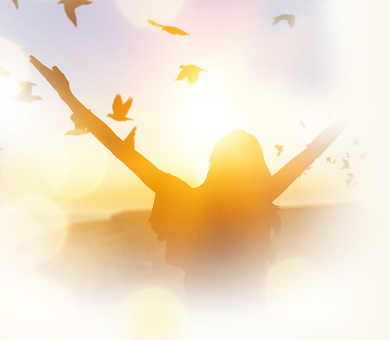 Mujer con los brazos en alto, mirando el ocaso mientras aves sobrevuelan su cabeza.